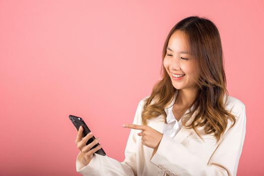 Portrait Asian happy beautiful young woman smile making finger pointing on screen smartphone in hand shopping online application looking to screen, studio shot isolated pink background with copy space