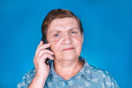 Elderly pensioner woman talking with mobile device on blue studio wall. Grandmother smiling. Technology, old people concept. High quality photo