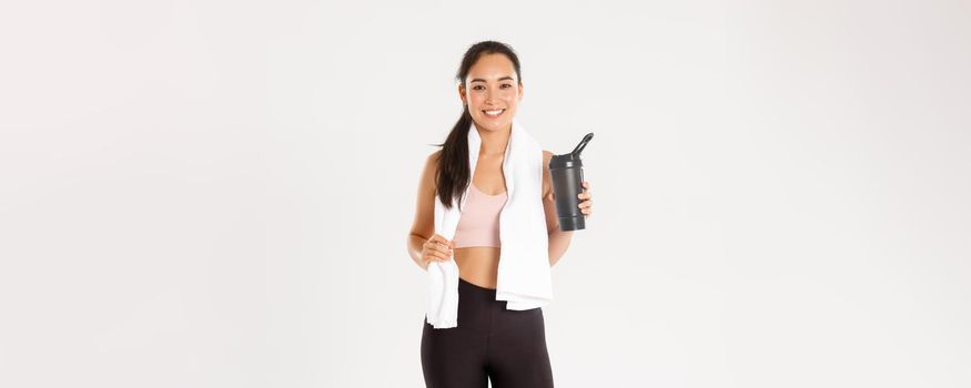 Sport, wellbeing and active lifestyle concept. Portrait of satisfied attractive asian fitness girl with cute smile, looking pleased wiping sweat with towel and drinking water after workout.
