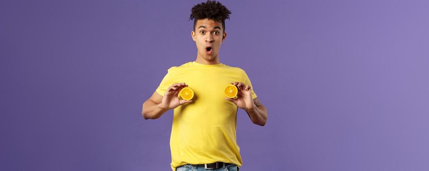 Holidays, vitamins and vacation concept. Portrait of funny and cute young 25s man fool around, showing breast with pieces of oranges over chest, look ashamed or shocked, purple background.