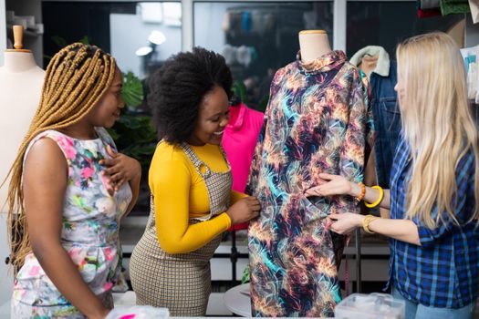 A clothing designer shows a dress from her new collection to two interested clients. Two African women and a European woman in the tailor's workshop.