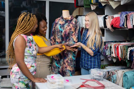 A clothing designer shows a dress from her new collection to two interested clients. Two African women and a European woman in the tailor's workshop.