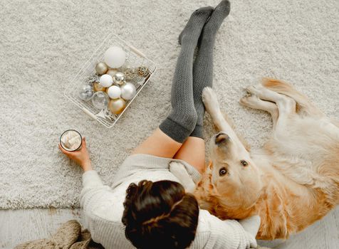 Girl holding mug with cocoa and marshmallow and golden retriever dog lying close to her in festive holidays time. Young woman with hot beverage and pet doggy at home