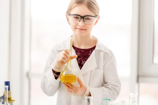 Smart girl during scientific chemistry experiment wearing protection glasses, holding bottle with orange liquid. Schoolgirl with chemical equipment on school lesson portrait