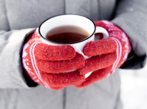 Hands wearing red knitted gloves holding mug with hot beverage cocoa in winter snow time outdoors. Person with warm tea coffee in cup