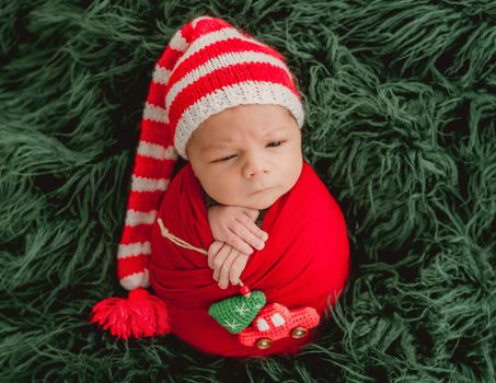 Newborn baby boy swaddled in fabric and wearing knitted Santa hat lying in basin decorated with Christmas tree. Cute infant child kid New Year studio portrait