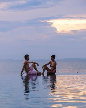 couple men and women relaxing on the edge of a pool during sunset by ocean at a luxury vacation. Men and women watching the sunset in the swimming pool