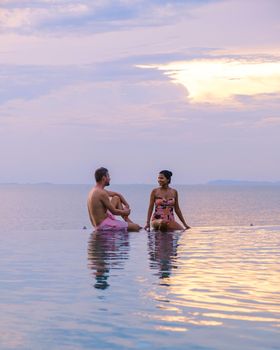 couple men and women relaxing on the edge of a pool during sunset by ocean at a luxury vacation. Men and women watching the sunset in the swimming pool