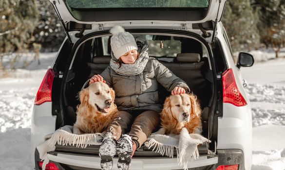 Girl sitting in car trunk with two golden retriever dogs in winter time and looking at camera. Young woman with doggy pet labradors in auto transportation outdoors