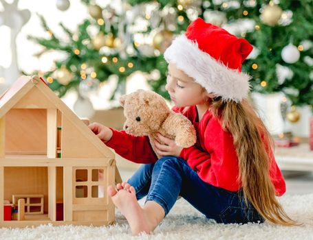 Little girl child wearing Santa hat in Christmas time sitting on floor and playing with wooden toy house and teddy bear at decorated New Year home with festive tree. Cute kid in Xmas time