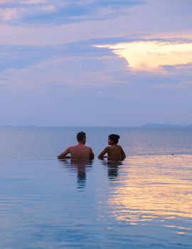 couple men and women relaxing on the edge of a pool during sunset by ocean at a luxury vacation. Men and women watching the sunset in the swimming pool