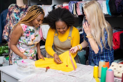Creative dark-skinned girls and one European woman match buttons to a selected sewing fabric. Two African girls and a European woman in a sewing studio.