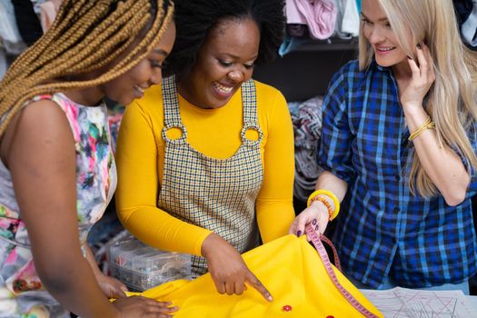 Creative dark-skinned girls and one European woman match buttons to a selected sewing fabric. Two African girls and a European woman in a sewing studio.