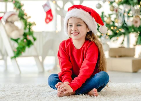 Little girl child wearing Santa hat in Christmas time sitting and smiling at decorated New Year home with festive tree. Cute kid in Xmas time