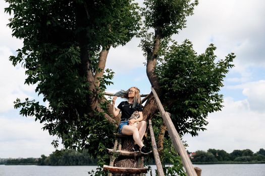 A young woman with a three-month-old Scottish Straight kitten sits on a beautiful green tree above the lake in summer. Walk, rest with a pet. The blonde is wearing shorts, a black T-shirt and glasses.