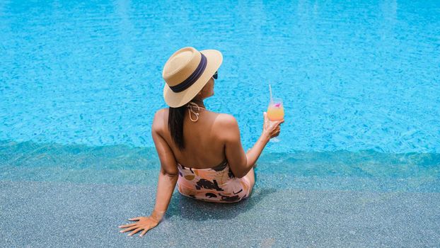 Asian women with a hat relaxing in the swimming pool with a cocktail in hand, women swimming pool banner holiday vacation concept, Asian women in blue swimming pool luxury vacation