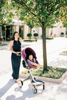 Mom in a long dress stands with a stroller with a baby under a tree. High quality photo
