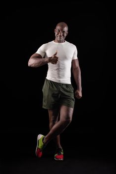 African American man standing looking at camera with thumb up and one leg crossed. Muscular adult male in sportswear in a studio with black background.