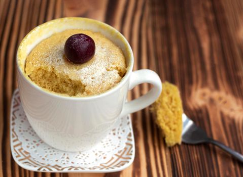 homemade cupcake in a mug cooked in the microwave. Decorated with cherries and icing sugar. without one piece. Part of the biscuit on a fork lies on a wooden table. Place for text. Cherry on the cake