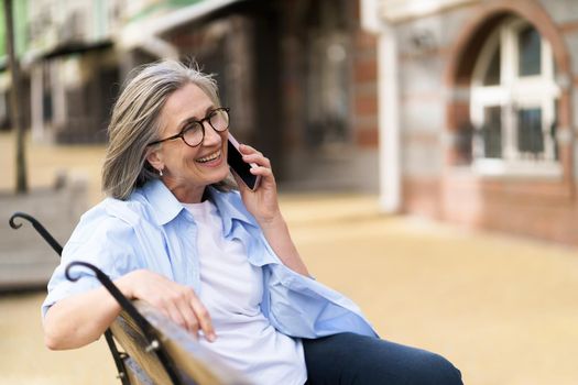 Mature grey hair woman smiling talking on the phone sitting on the bench enjoying summer time at the streets of old European town. Mature woman answering call outdoors.