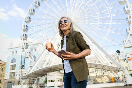 Happy grey haired European woman enjoying traveling, holding phone, smartphone while having juice on the go using plastic cup wearing sunglasses and green shirt in city and ferris wheel on background.