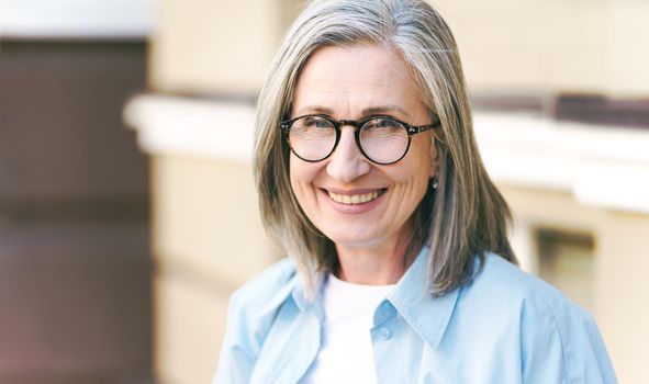 Charming grey hair mature woman wearing eye glasses standing outdoors enjoying vacation traveling around world at retirement. Mature woman with perfect skin wearing blue shirt.