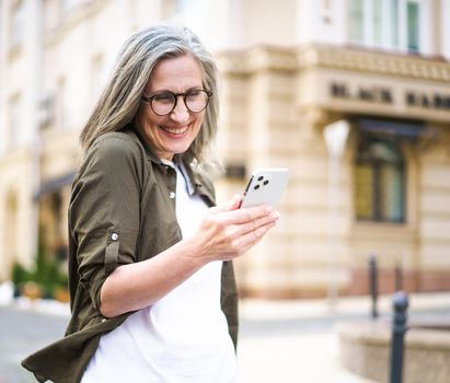 Mature beautiful grey hair woman looking at smartphone walking at the streets of old European town. Senior businesswoman read text message, answering video call standing outdoors. Selective focus.