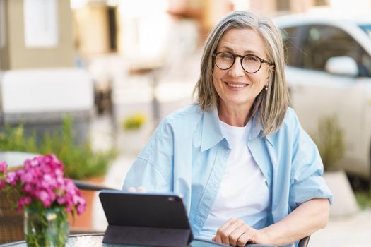 Mature grey haired European woman using digital tablet sitting at table of a small street cafe. Mature silver hair woman working outdoors city cafe, wearing white t-shirt and blue shirt.