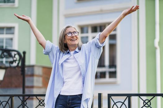 Happy excited mature woman with hands lifted up wearing glasses standing outdoors with urban city background. Senior woman praising god outdoors. Giving thanks to God mature woman.