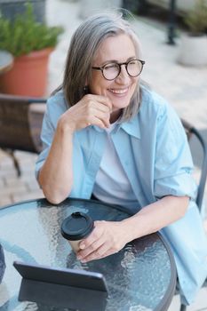 Grey haired mature European smiling woman drink coffee using digital tablet sitting at table of street cafe. Mature silver hair woman working outdoors city cafe, wearing white t-shirt and blue shirt.