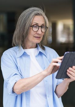 Interested in reading news or social media mature grey hair woman holding digital tablet sitting standing outdoors at the city streets. Mature woman answering call outdoors.
