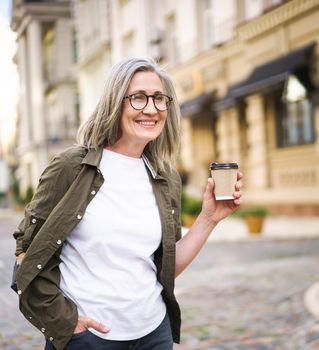 Happy mature businesswoman with disposable cup of hot coffee in hand. Charming senior woman traveling in Europe enjoying cup of tea, coffee. Grey haired woman in casual with backpack.