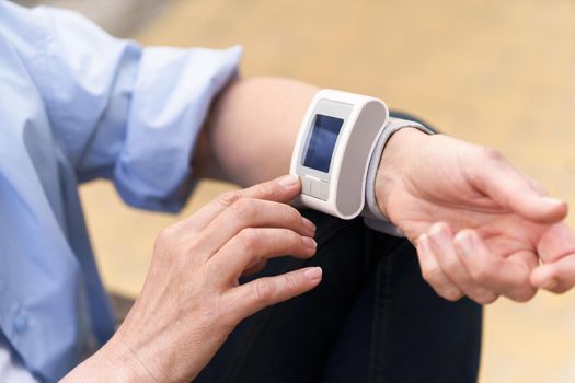 Mature woman use arm pulse, blood pressure monitor sitting outside street and making health examine. Senior woman various situation outdoors. No face visible.