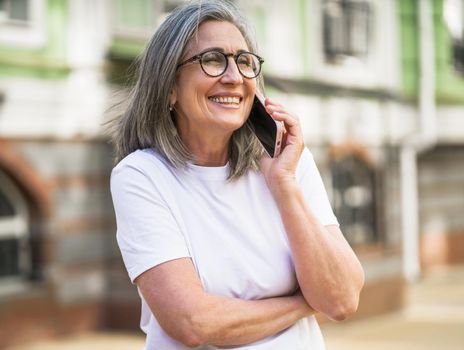 Mature grey hair business woman in eye glasses talking on the phone standing outdoors of the streets of old urban city wearing white t-shirt. Silver hair woman outdoors.