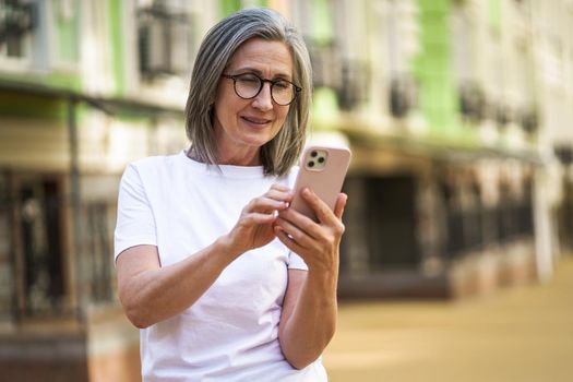 Mature woman with modern technologies. Senior grey hair lady holding smartphone standing outdoor at the streets of old town. Mature woman read text message standing or walking outdoors.