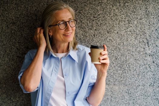 Enjoying her coffee mature grey hair woman standing leaned on marble wall smiling looking sideways away fixing her hair. Standing outdoors drinking coffee positive gorgeous woman in casual blue shirt.