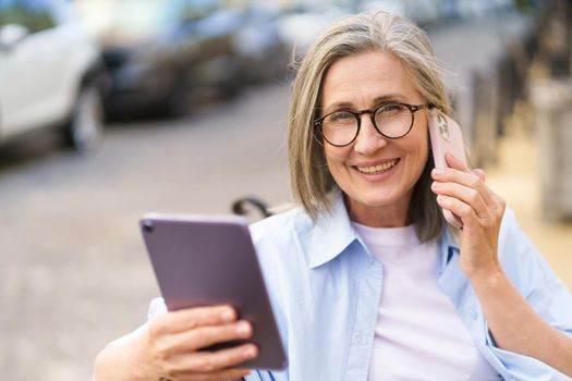 Reading updates on digital tablet mature grey hair woman talking on the phone sitting on the bench at the streets of old European city. Mature woman answering call outdoors.