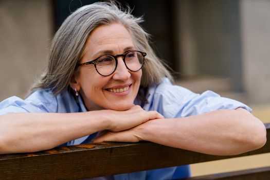 Leaned on the bench happy mature grey hair woman smiling sitting enjoying summer time at the streets of old European town. Mature people leisure retirement life.