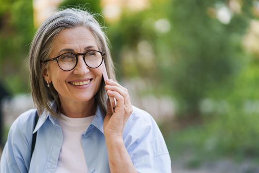 Senior grey hair woman in eye glasses smiling talking on the phone standing outdoors garden or park background wearing blue shirt. Silver hair woman outdoors.