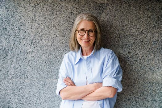 Beautiful mature woman with grey hair leaned on marble wall standing with folded arms outdoors smiling looking at camera. Standing outdoors positive gorgeous woman in casual blue shirt.