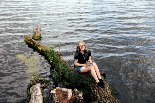 Young blonde woman with blue hair, wearing glasses, denim shorts and a black T-shirt. Sitting on a floating tree near the lake with a three-month-old kitten breed Scottish Straight.