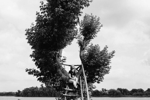 A young woman with a three-month-old Scottish Straight kitten sits on a beautiful green tree above the lake in summer. Walk, rest with a pet. The blonde is wearing short shorts, a T-shirt and glasses.