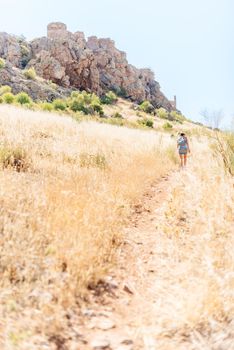 Vertical photo of a woman in a hat and summer clothes climbing a hill to a romanesque castle on top in Toledo, Spain