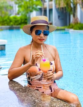 Asian women with a hat relaxing in the swimming pool with a cocktail in hand, women swimming pool banner holiday vacation concept, Asian women in blue swimming pool luxury vacation