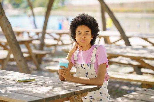 Wistful African American female with takeaway coffee looking away while sitting at wooden table in park on sunny summer day
