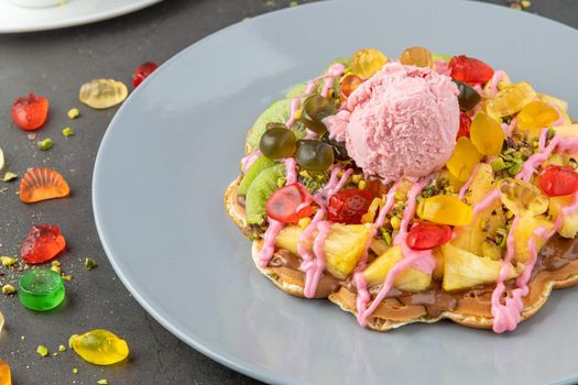 Heart waffle with kiwi and pineapple with gummy candy and ice cream on it.