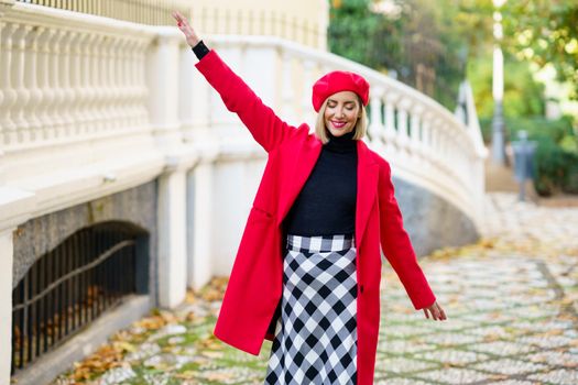 Positive female in red clothes with beret strolling with raised arm and closed eyes on street along white fence in city