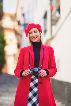 Self assured young female tourist, with blond hair in elegant outfit and beret with photo camera standing on narrow street and smiling at camera during sightseeing trip in old town