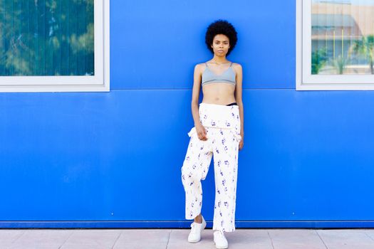 Full body of slim African American female in overall and bra looking at camera while standing on street near blue building
