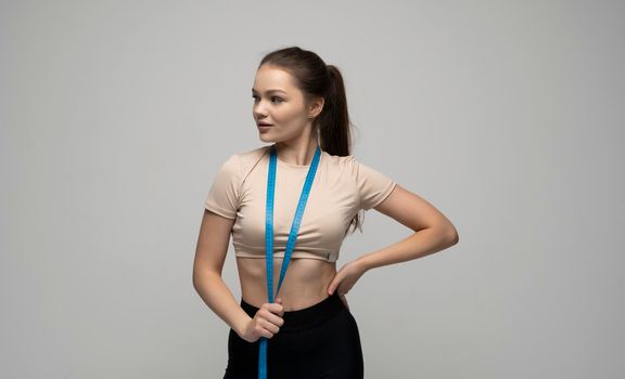 Portrait of happy brunette fitness girl in beige sportswear. Young smiling woman is measuring her body with tape measure and showing diet result